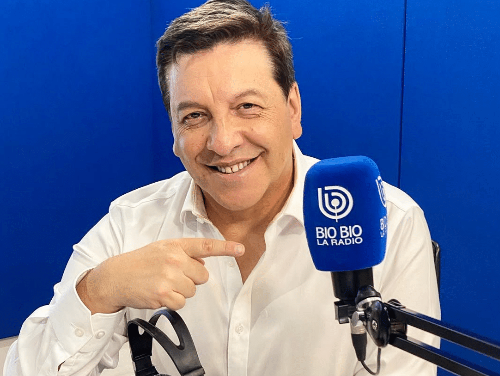Un hombre con camisa blanca sonríe y apunta hacia un micrófono azul de Bio Bio La Radio, frente a un fondo azul.