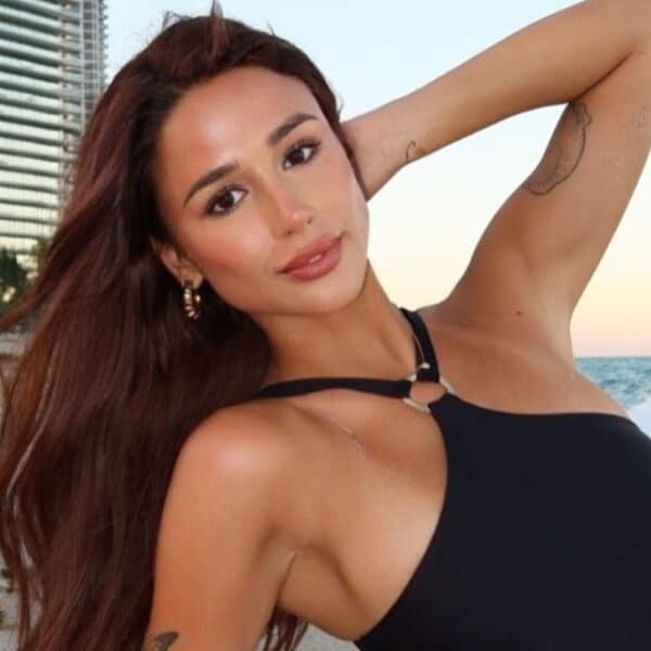 Mujer de cabello largo posa con un traje de baño negro en la orilla de una playa; detrás se ven el mar y un edificio.