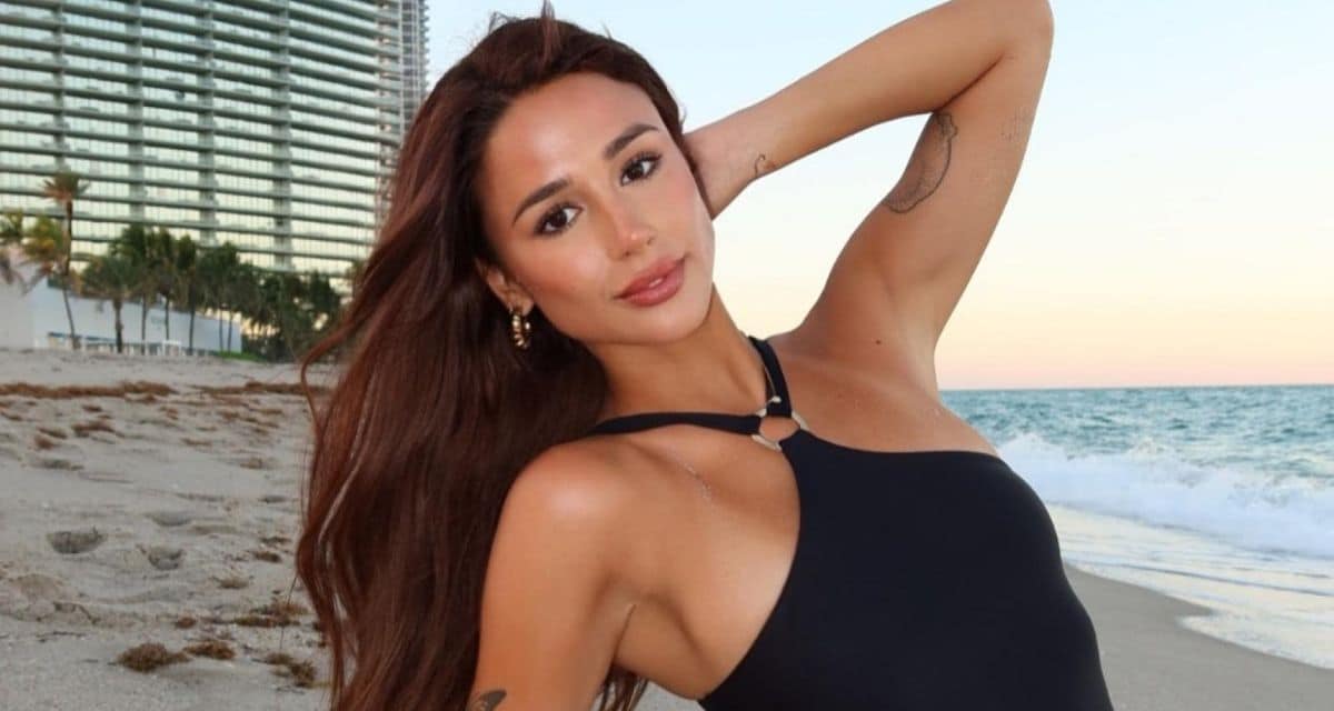 Mujer de cabello largo posa con un traje de baño negro en la orilla de una playa; detrás se ven el mar y un edificio.