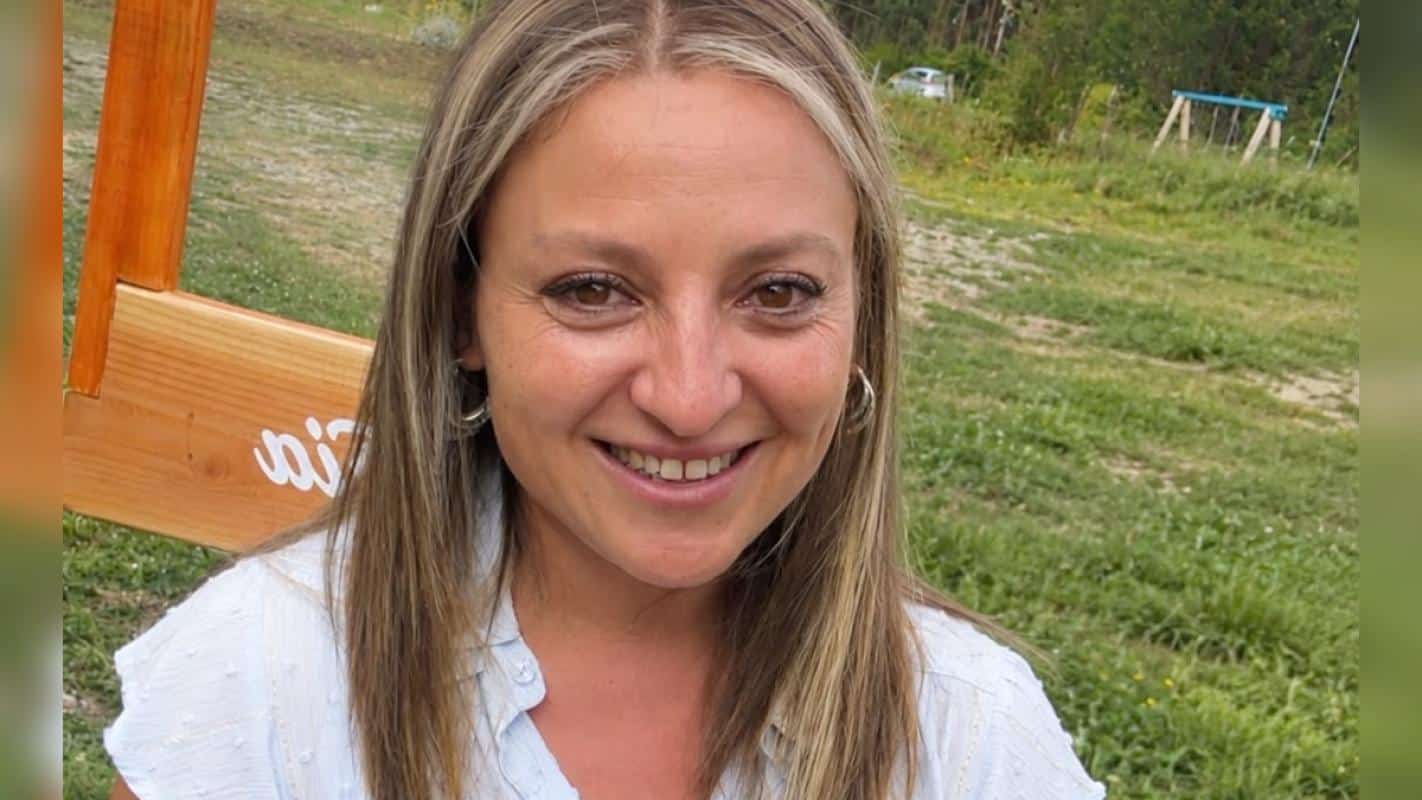 Mujer de cabello largo y claro sonríe a la cámara, con una pradera y una estructura de madera al fondo.