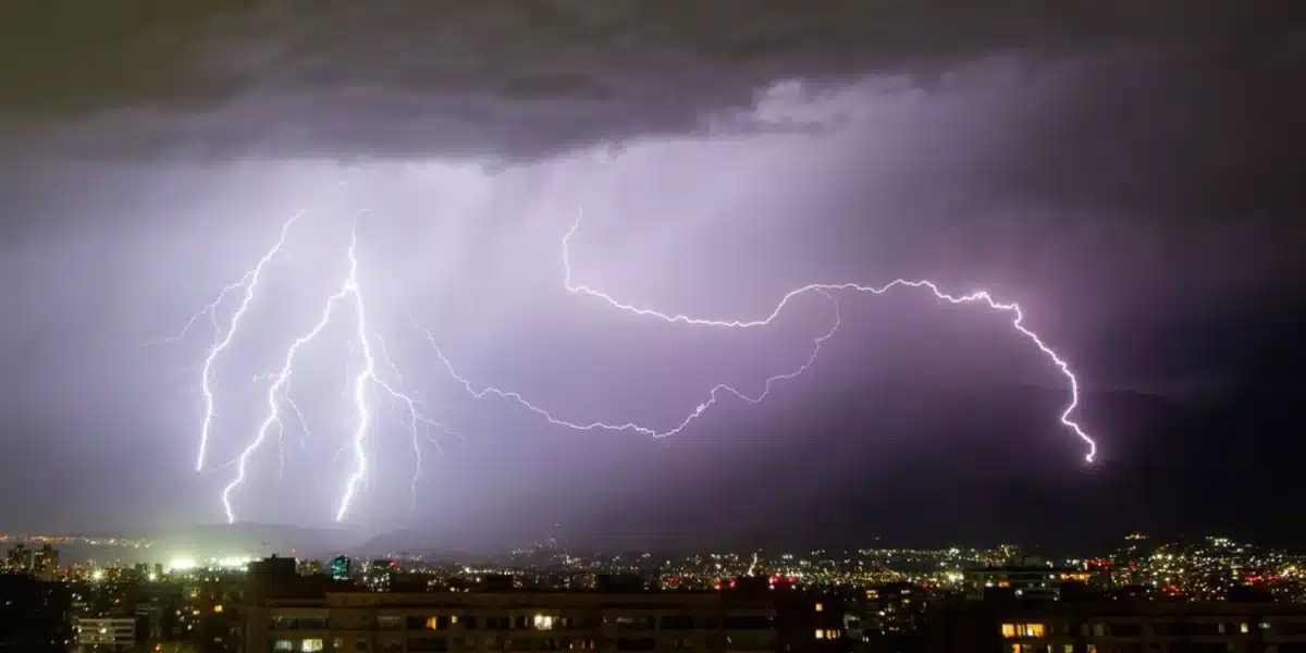 Varios relámpagos cruzan un cielo cubierto de nubes sobre una ciudad de noche, con edificios y luces en el horizonte.